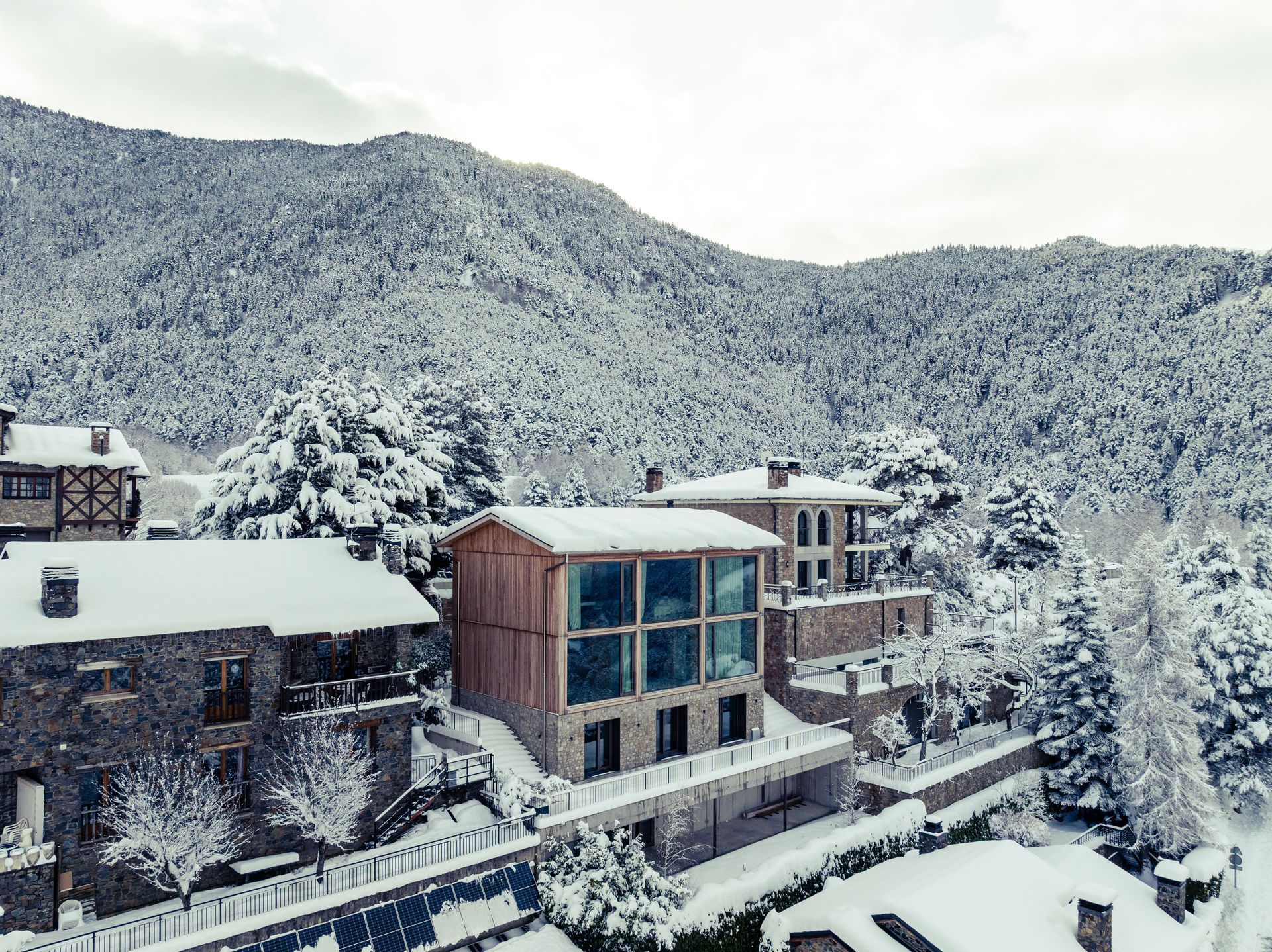 Poble de muntanya cobert de neu amb edificis moderns i arbres sota un vessant hivernal
