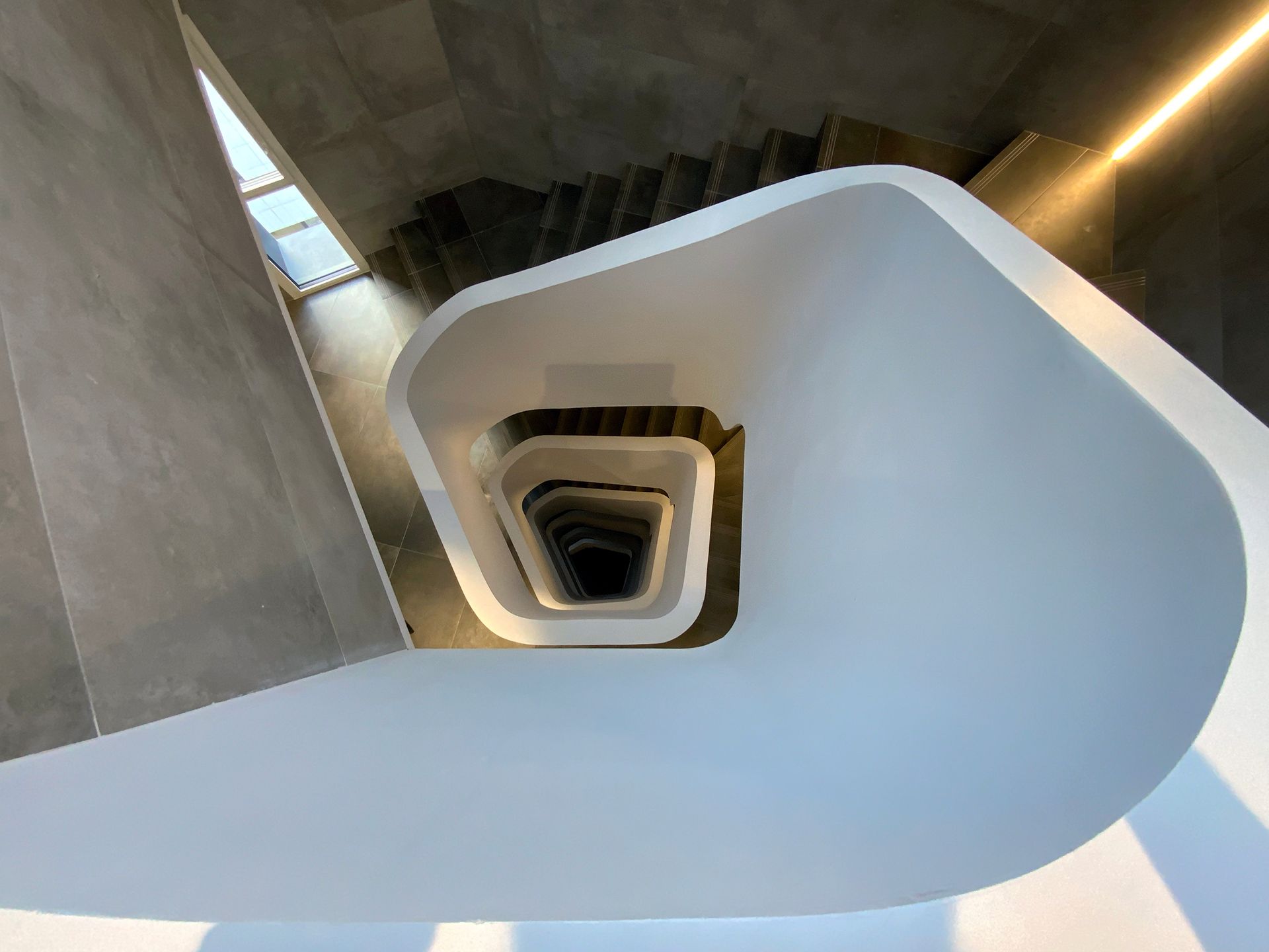 Escala interior de cargol amb barana blanca i parets de formigó, vista des de dalt mirant cap al centre.