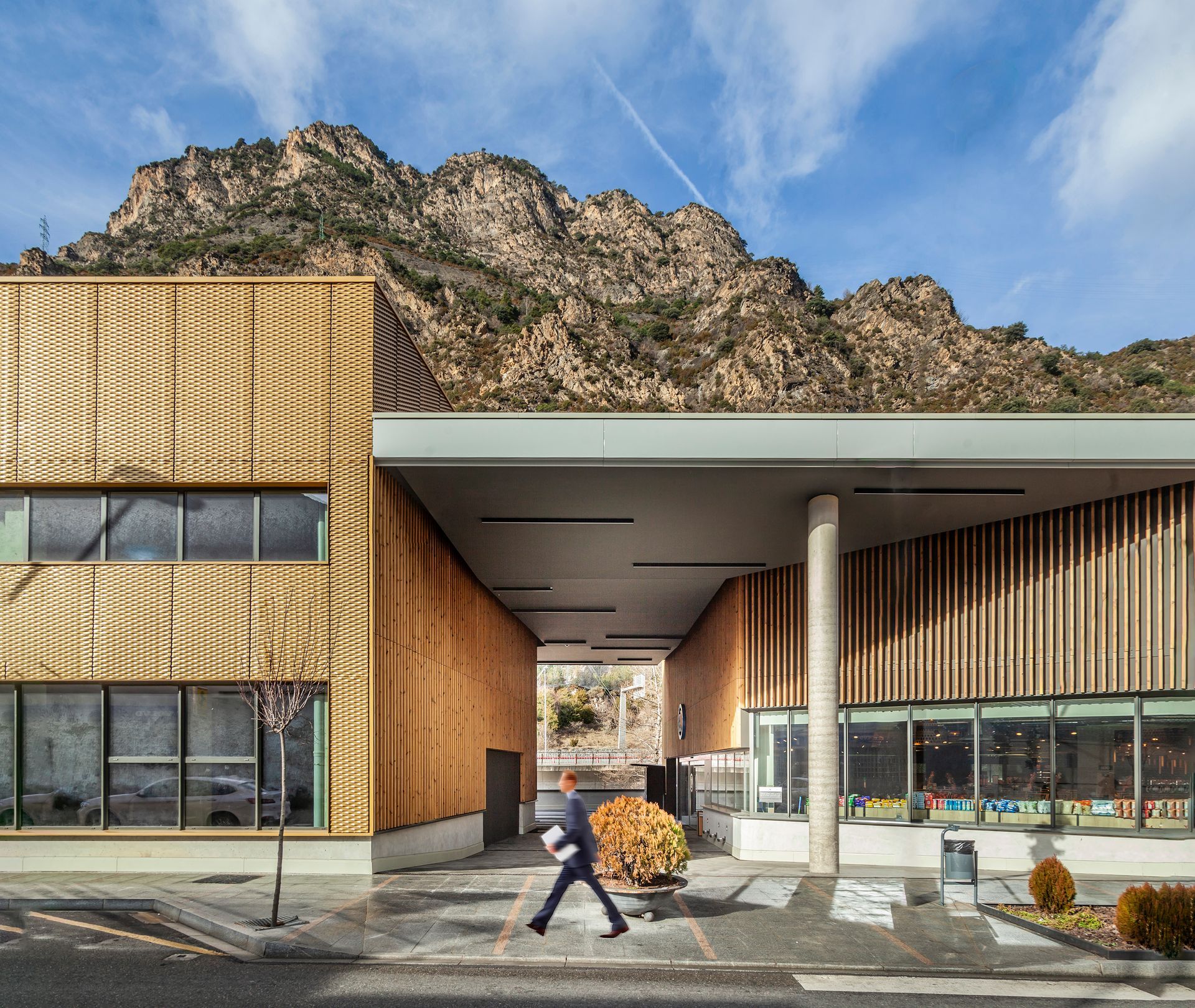 Entrada d'edifici modern amb façana de fusta i vidre, muntanyes al darrere i una persona caminant al davant.