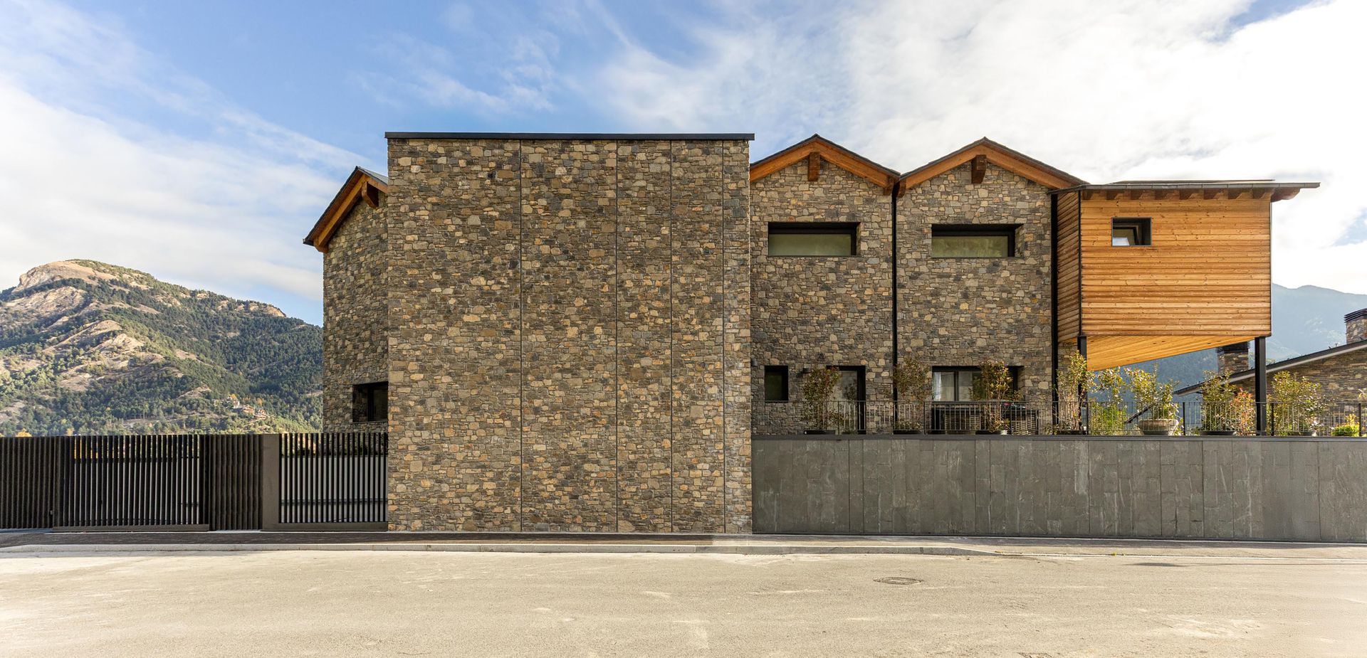 Casa moderna de pedra i fusta amb un teló de fons de muntanya sota un cel parcialment ennuvolat