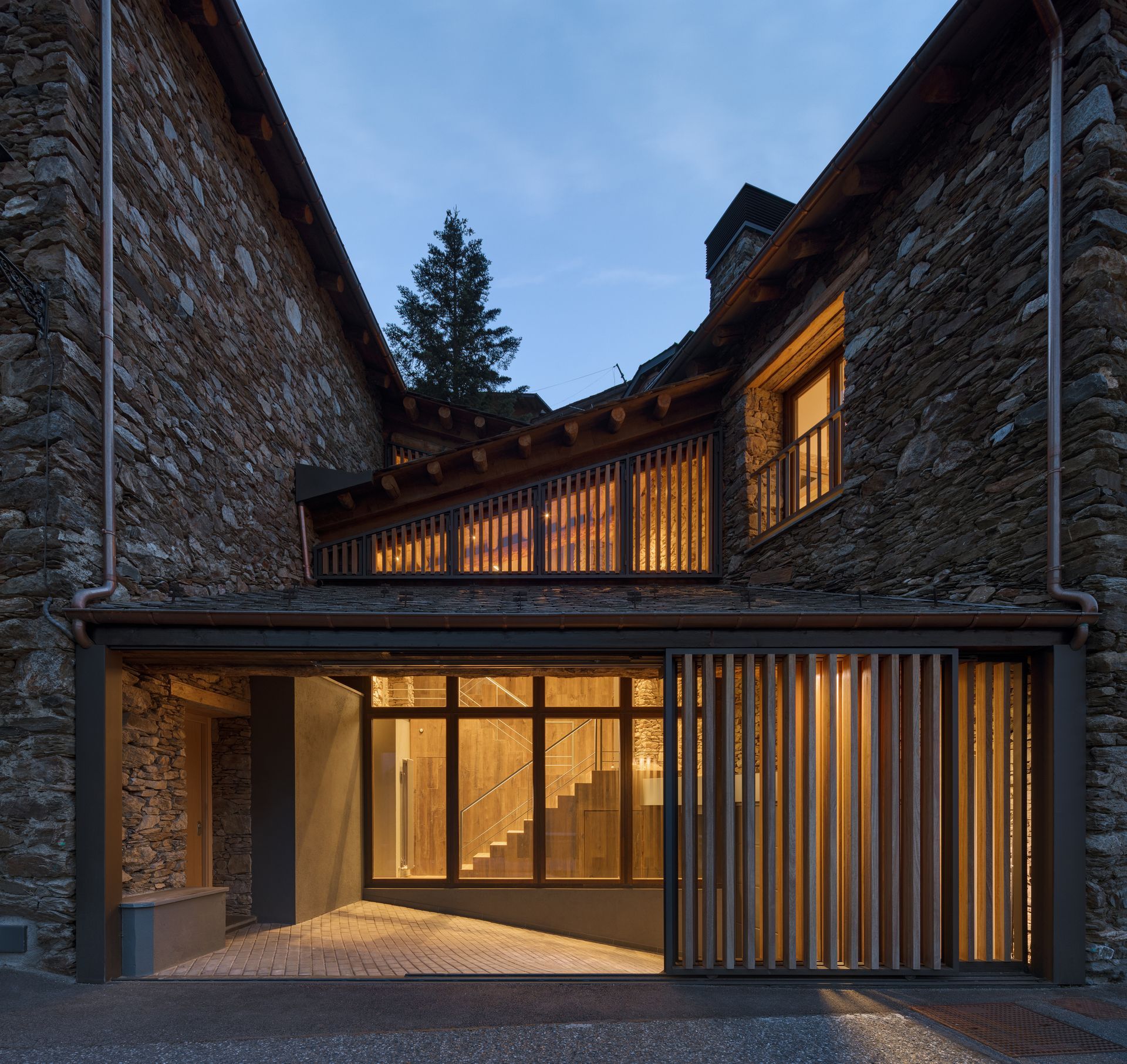 Exterior de casa de pedra moderna al capvespre amb finestres càlides i una elegant entrada de vidre i fusta