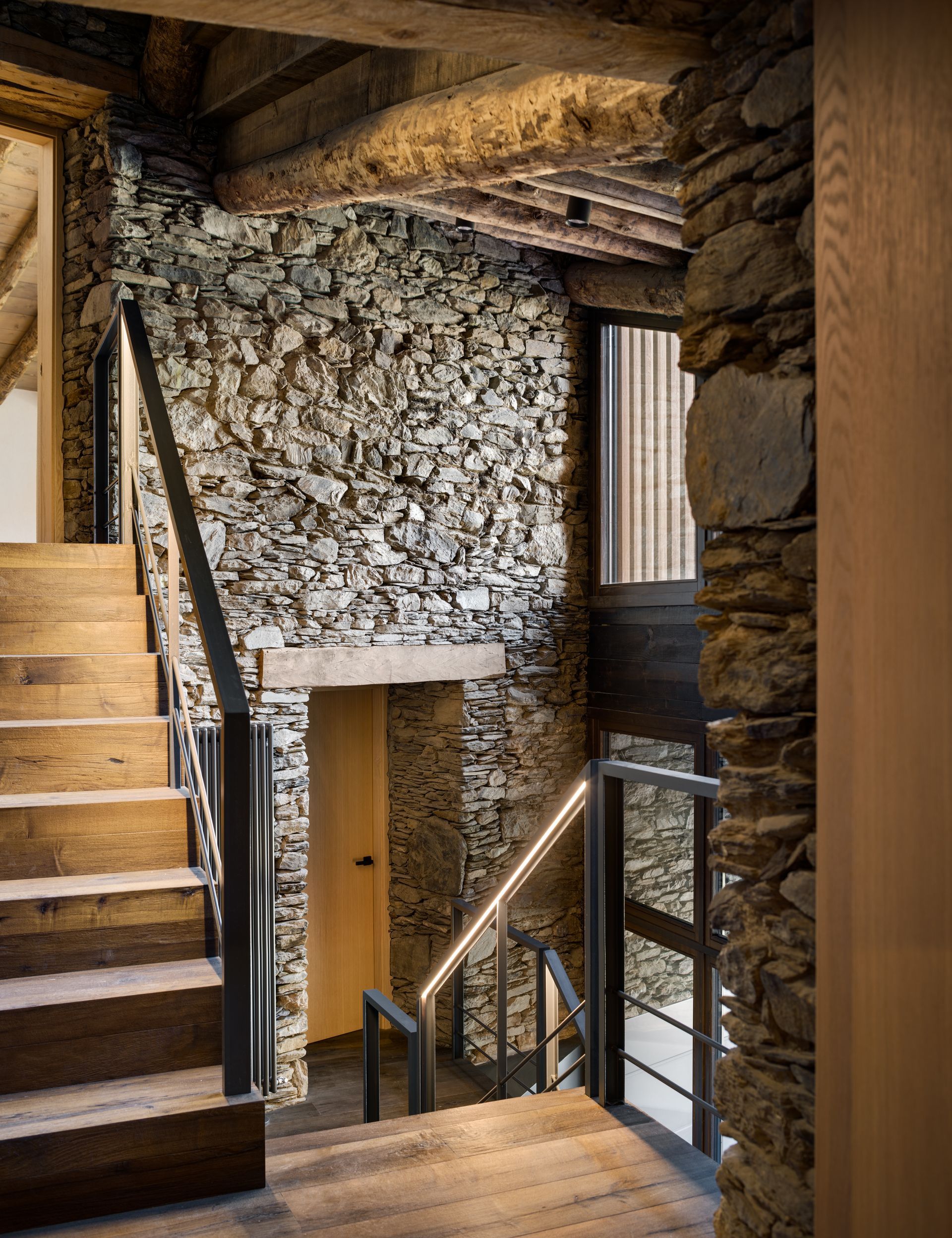 Escala rústica de pedra amb bigues de fusta, barana metàl·lica i finestres que deixen entrar la llum natural