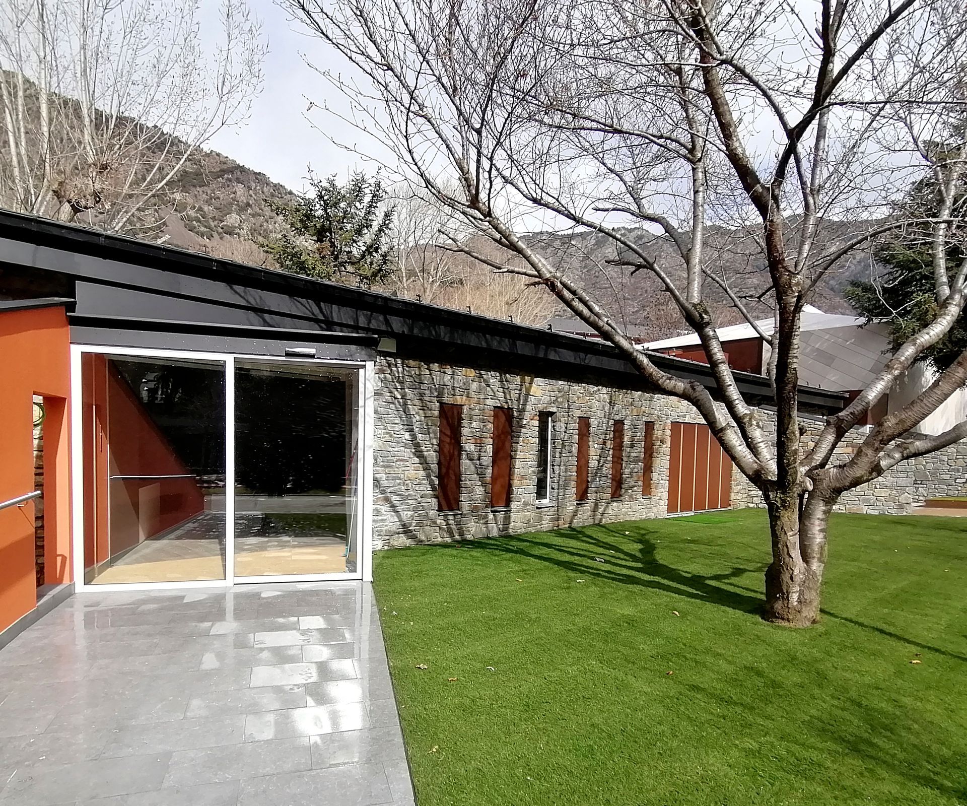 Edifici modern amb portes de vidre, parets taronges, gespa verda i un arbre sense fulles al davant