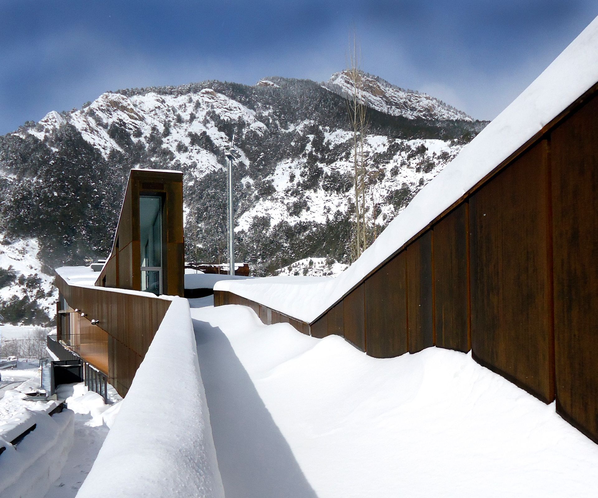 Passeig cobert de neu a la teulada al costat d'un edifici de fusta amb muntanyes al fons