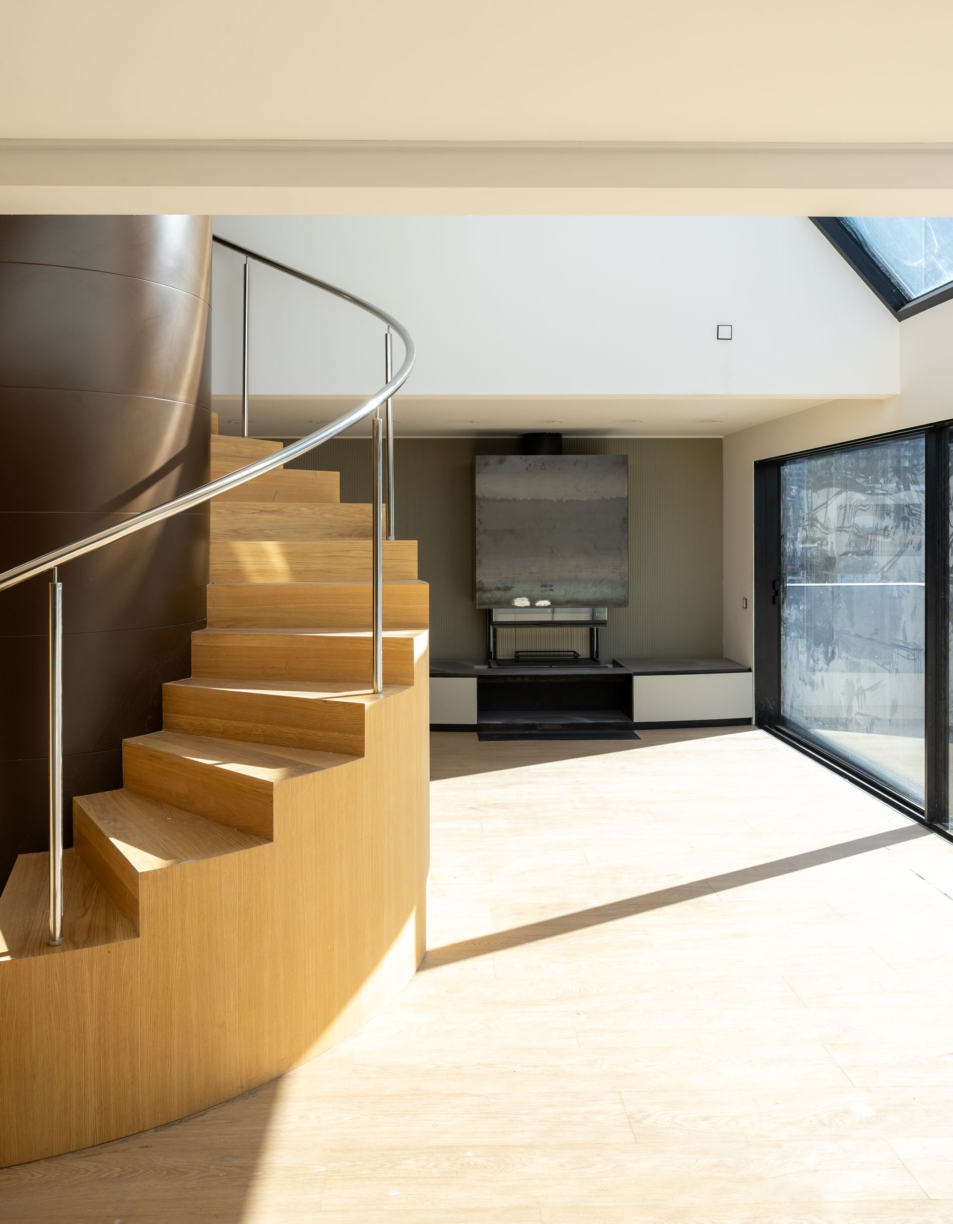 Interior modern amb escala de fusta corbada, sala d'estar oberta i llum solar brillant a través de grans portes de vidre