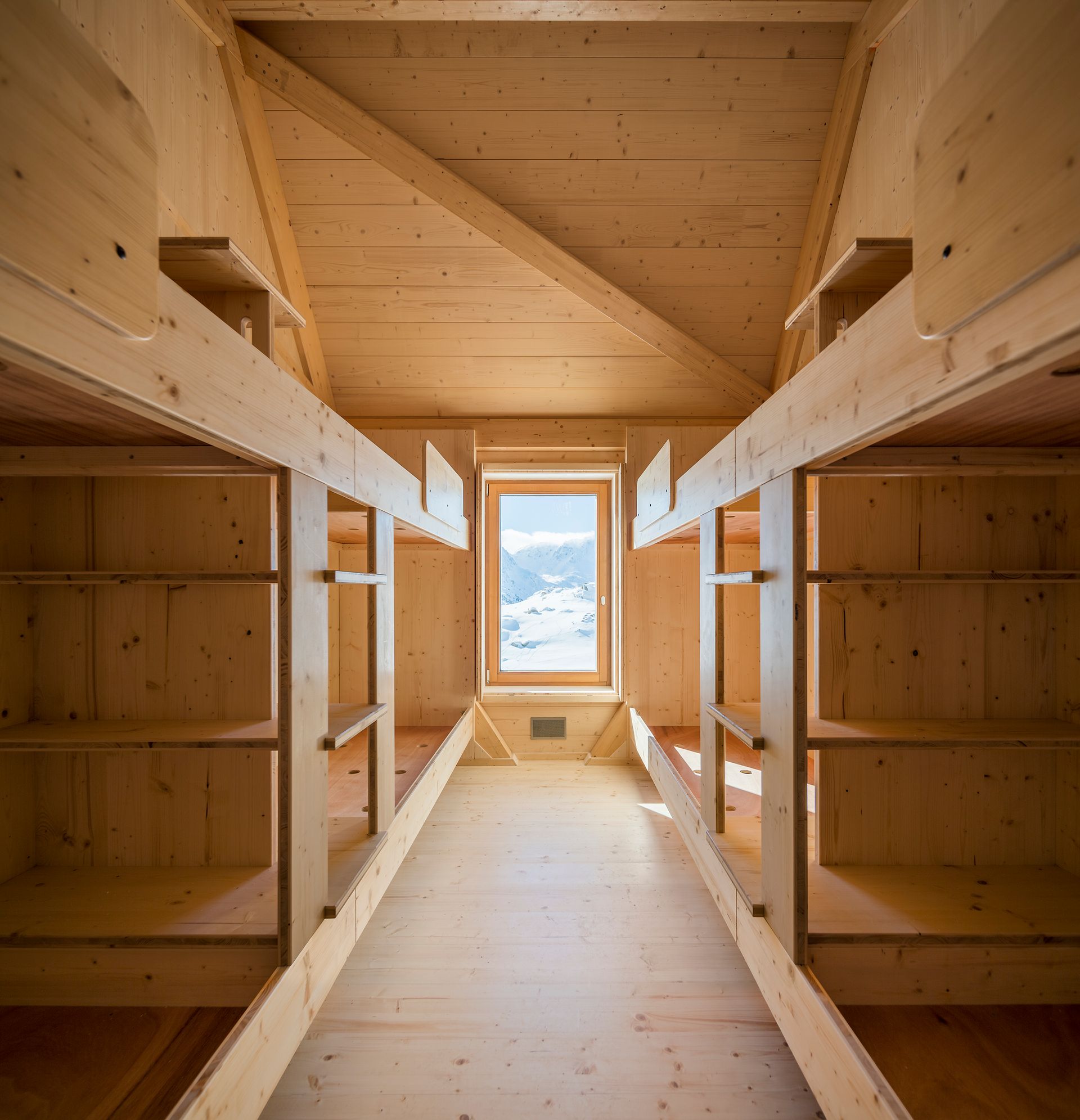Interior d'una habitació amb lliteres de fusta amb un passadís central i una finestra que emmarca muntanyes nevades.
