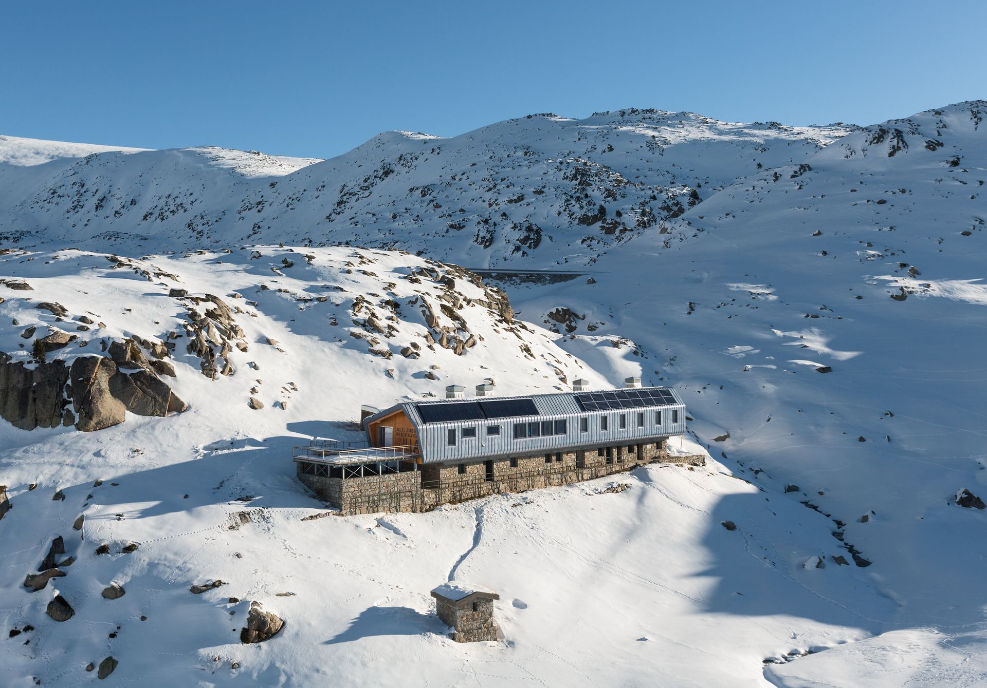 Paisatge de muntanya cobert de neu amb un petit refugi a la base de vessants escarpats i rocosos.