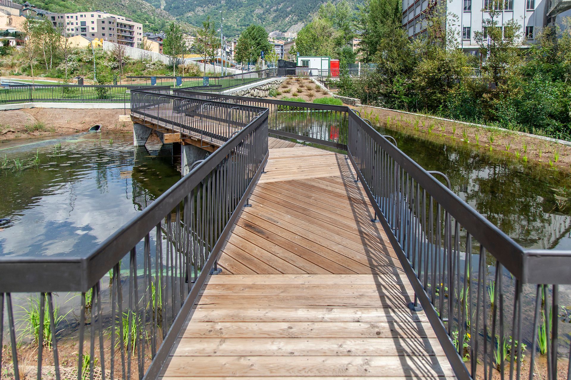 Pont de vianants de fusta sobre un estany amb baranes, envoltat de vegetació i edificis al vessant d'un turó