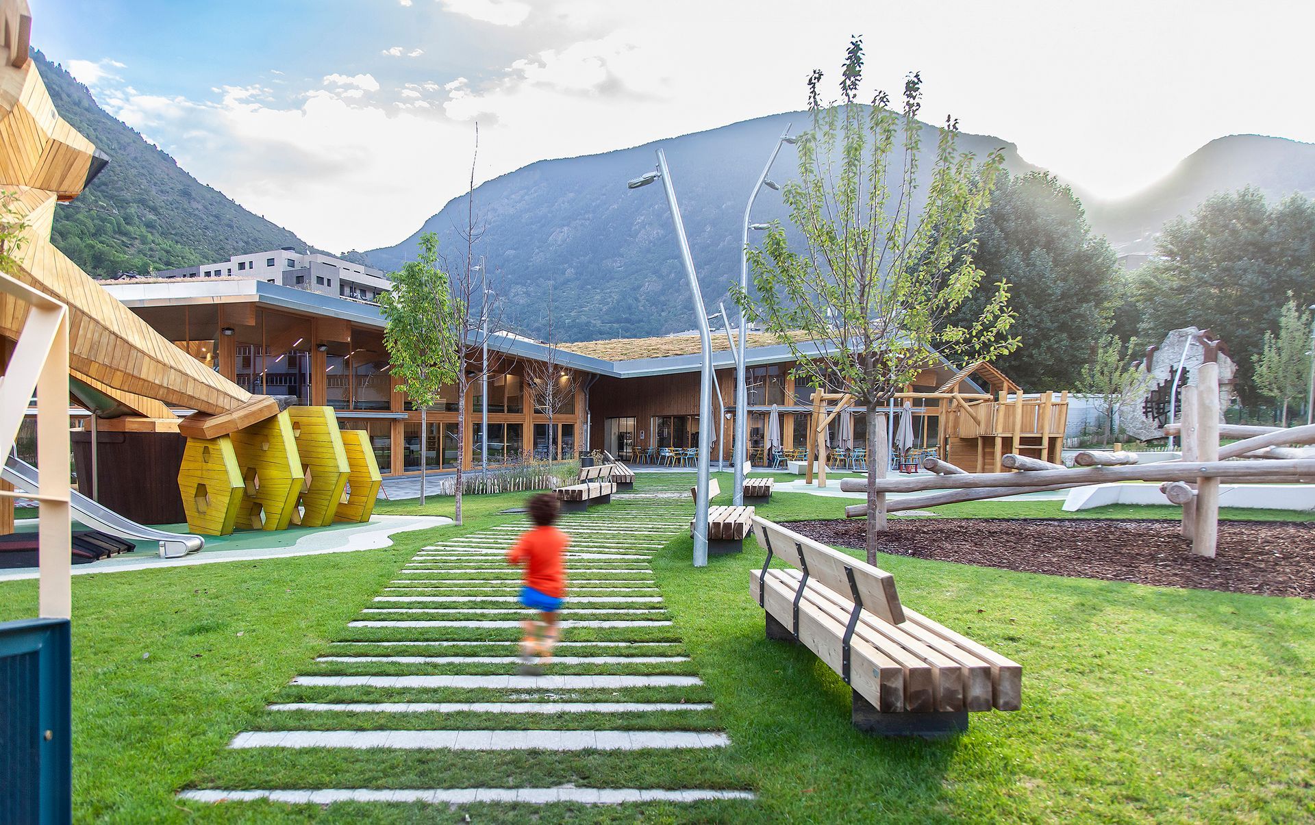 Nen caminant sobre pedres en un parc infantil amb gespa al costat d'un edifici modern de fusta i bancs
