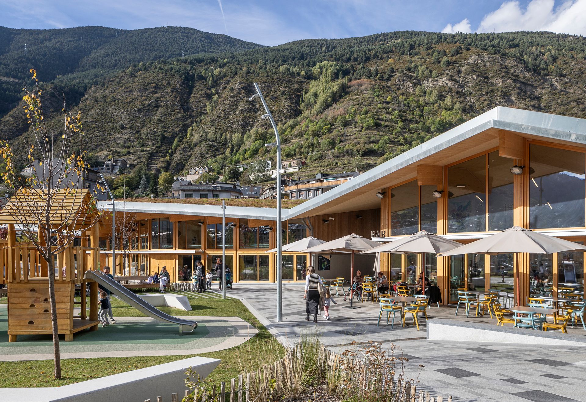 Pati d'un modern complex turístic de muntanya amb edificis de fusta, para-sols i un parc infantil contra turons rocosos