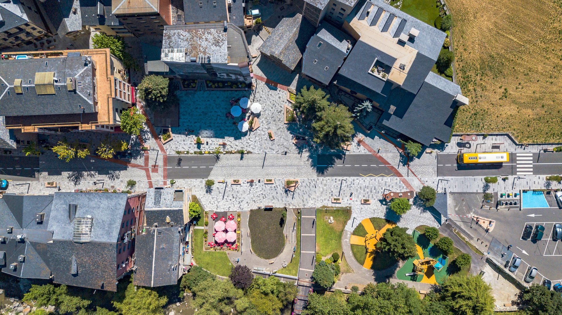 Vista aèria d'una petita plaça amb vianants, cotxes aparcats, para-sols de colors i edificis circumdants