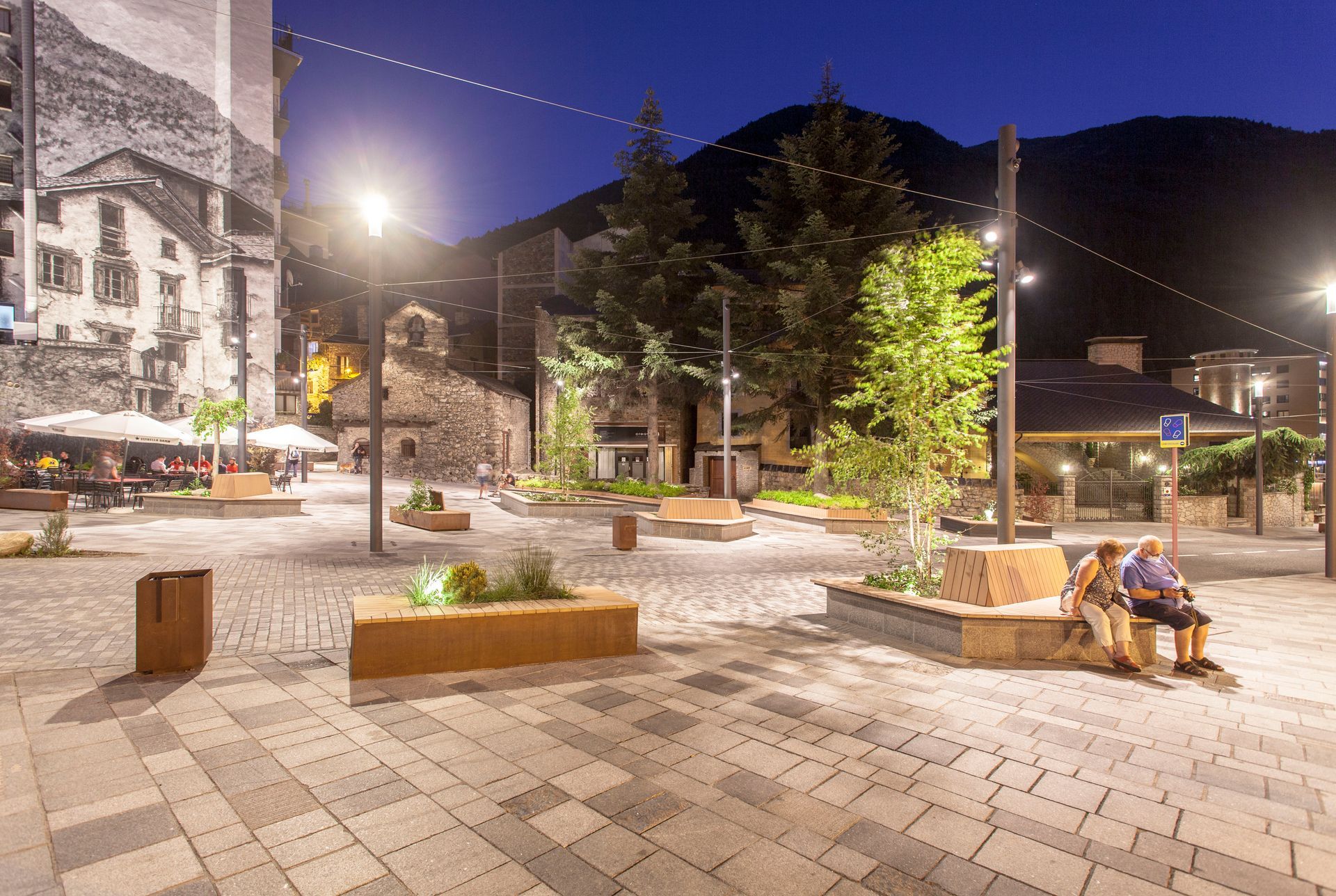 Plaça nocturna amb bancs, arbres, guirnaldes de llums i gent asseguda sota els càlids fanals del carrer.