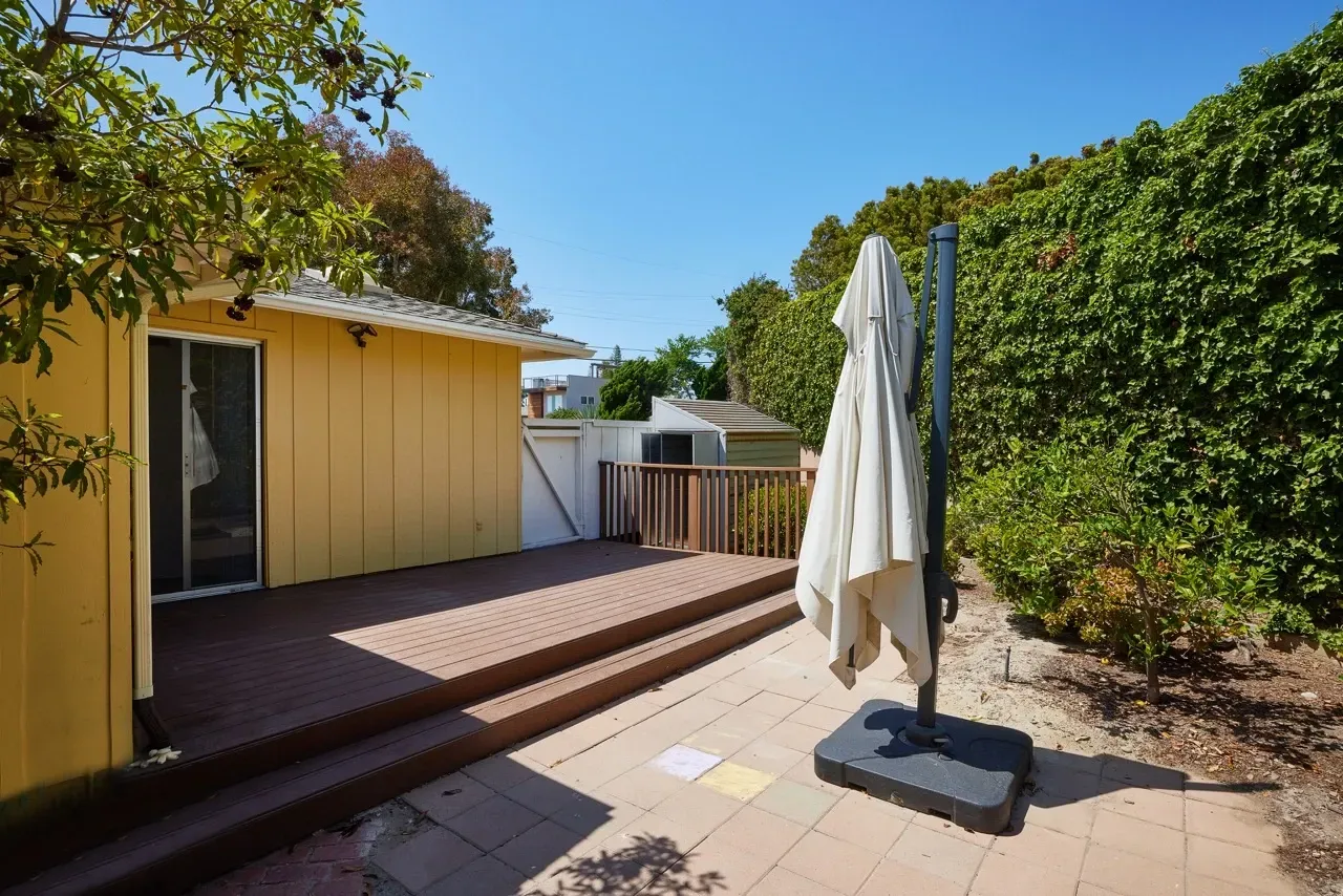 Private deck and patio area with a closed umbrella, surrounded by lush greenery and a vine-covered wall.