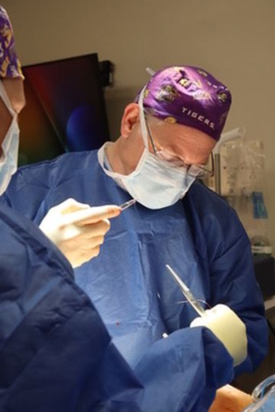 Surgeon in blue scrubs, mask, and purple Tigers cap, operating.