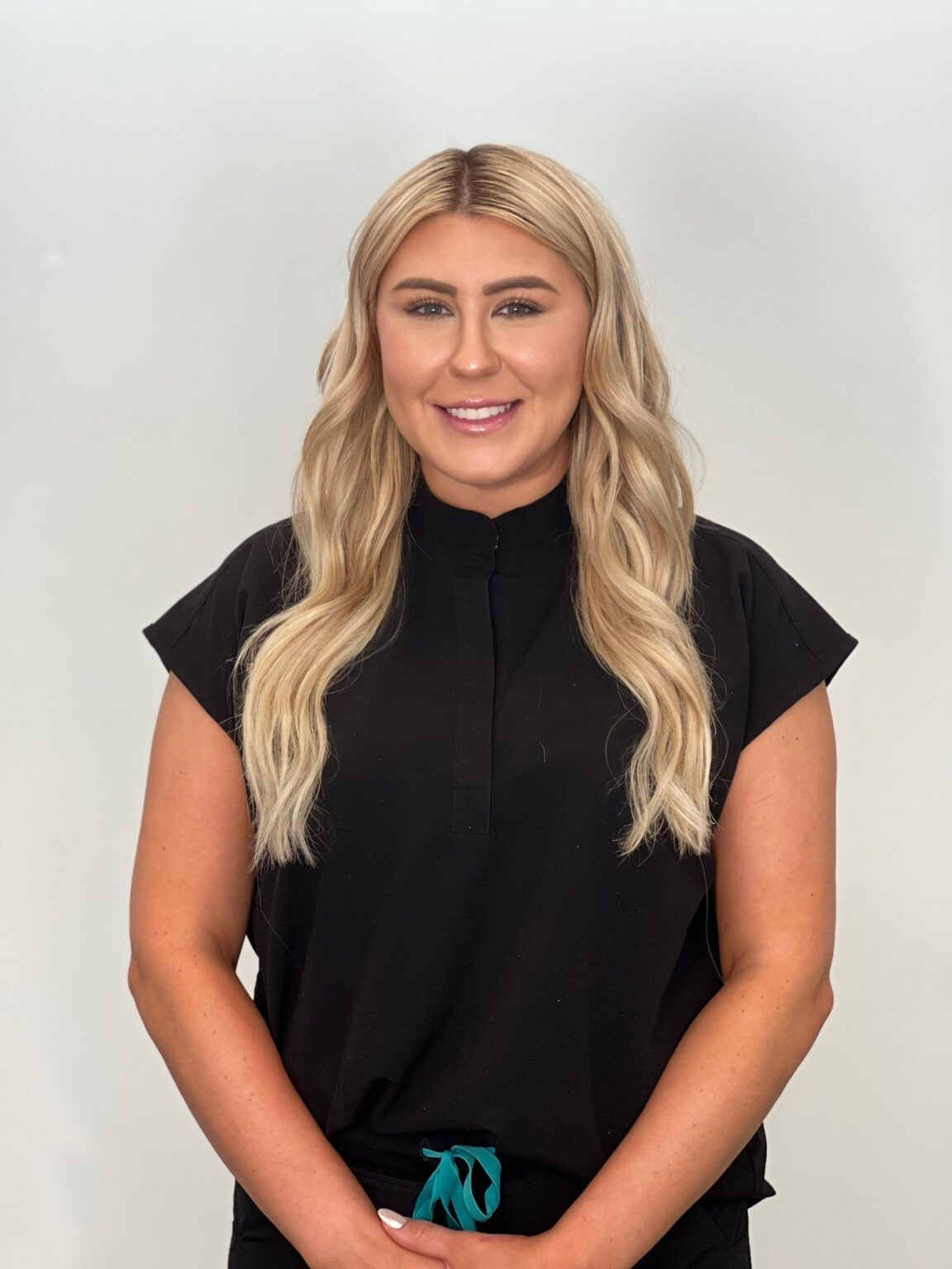 Woman with blonde wavy hair, wearing black top, smiling, posing against a white background.