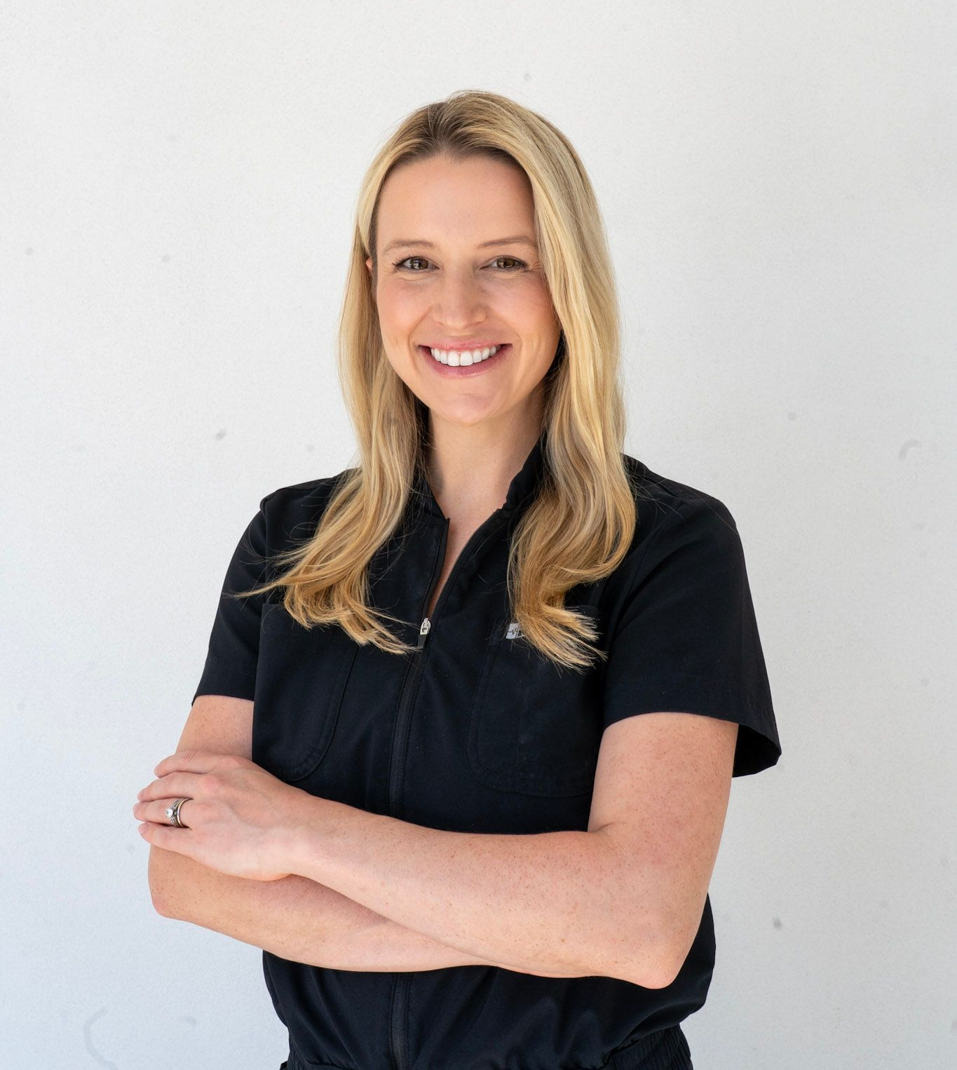Woman in black scrubs, smiling, hands on hips, standing in front of a white wall.