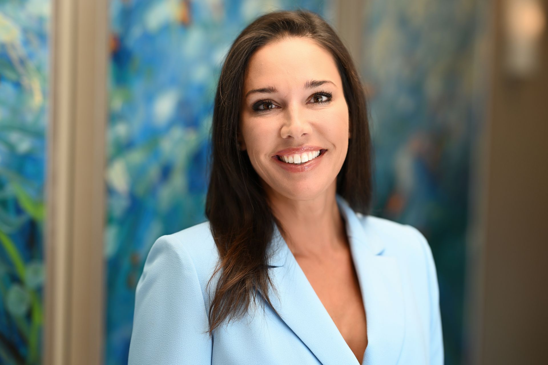 Woman in a light blue blazer smiles at the camera, standing in front of blue artwork.