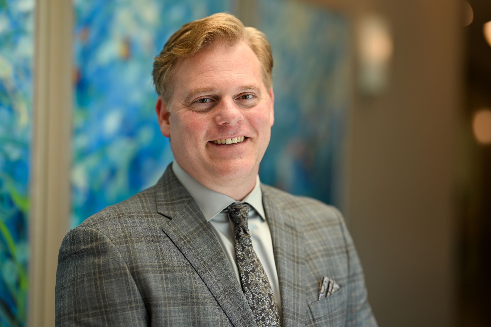 Man in plaid suit, smiling, in front of blue and white abstract art.