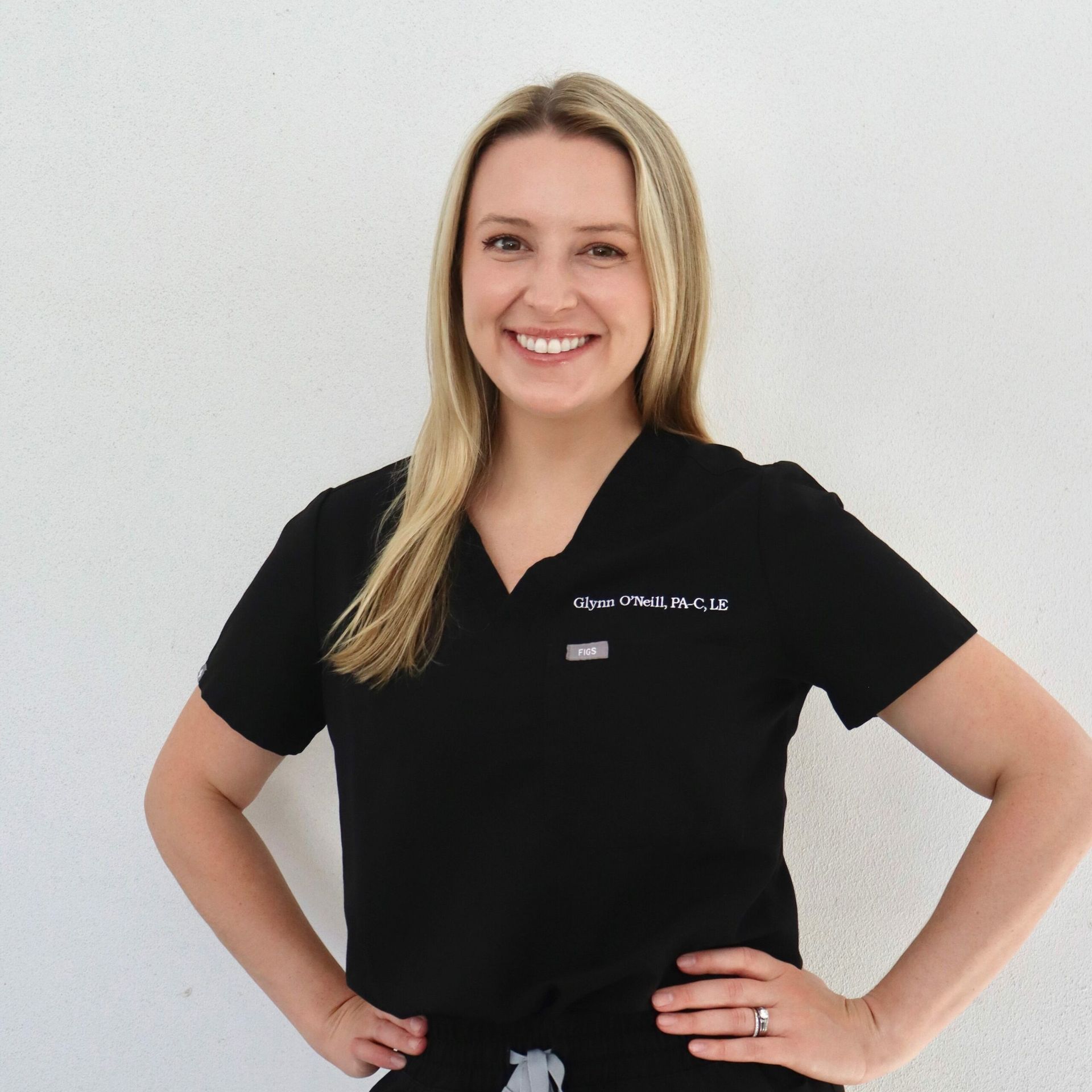 Woman in black scrubs smiles, hands on hips, against a white wall.