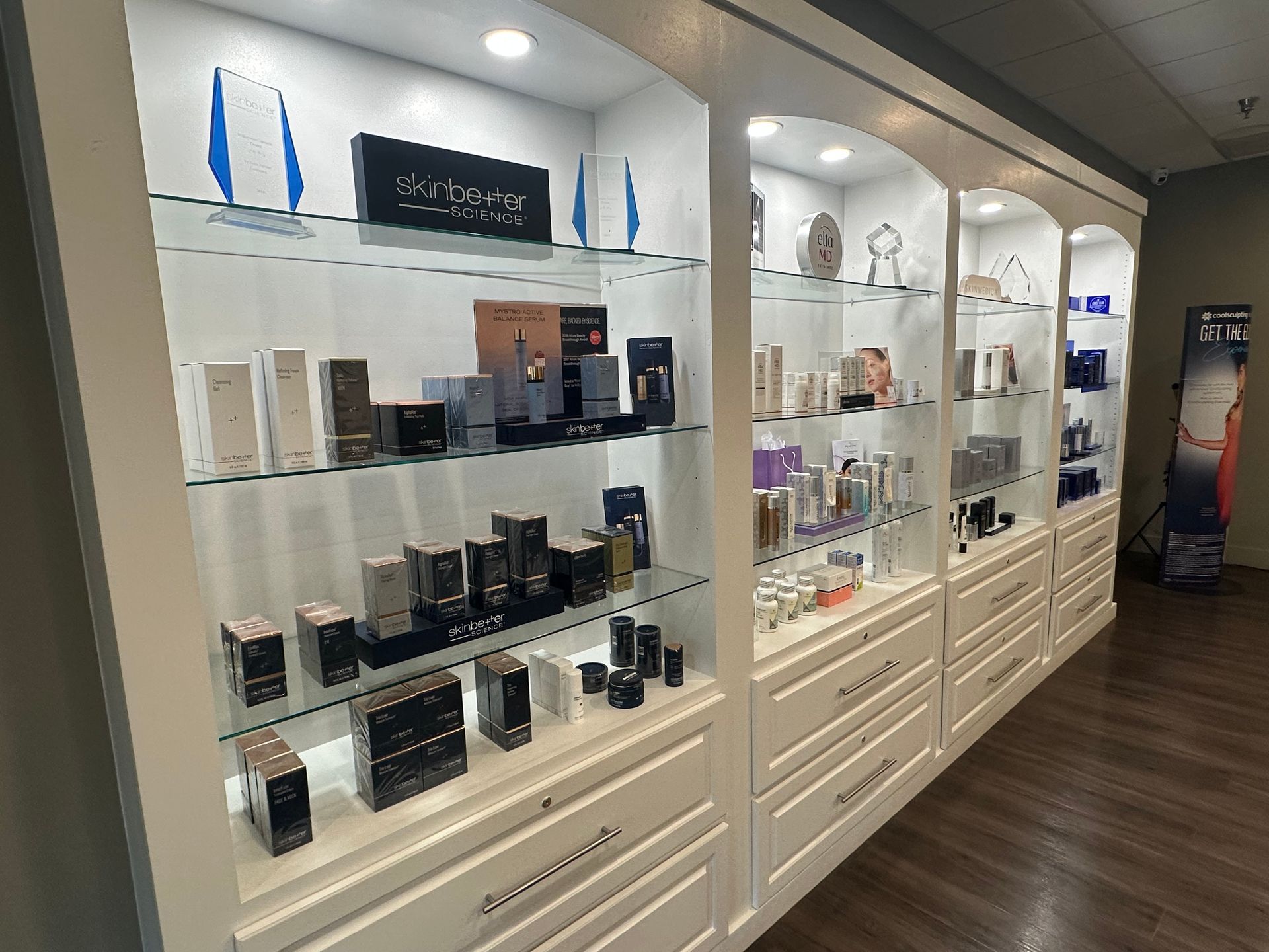 White display cabinets with skincare products, lit by spotlights. Wooden floor, neutral walls.