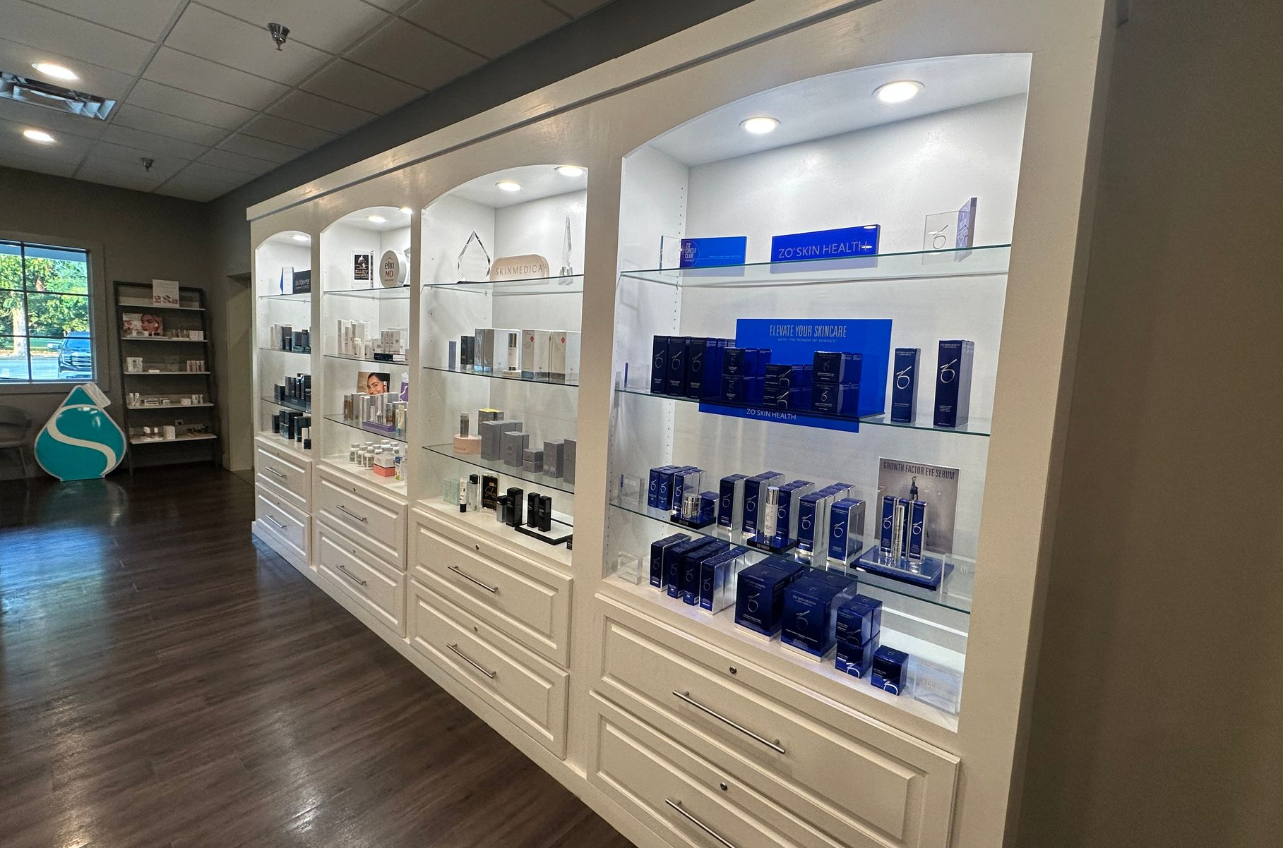 A well-lit display case with skincare products. White cabinets with glass shelves, dark wood floor.