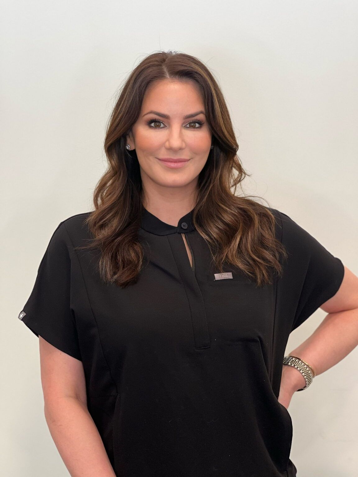 Woman with long brown hair, wearing a black shirt, smiles. Against a plain white wall.