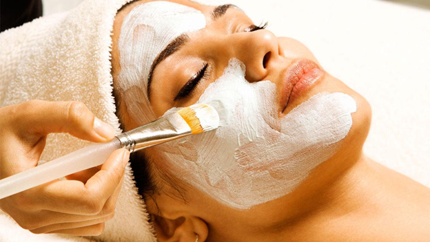 Woman receiving a facial mask application. A hand holds a brush, applying white cream. Eyes are closed.