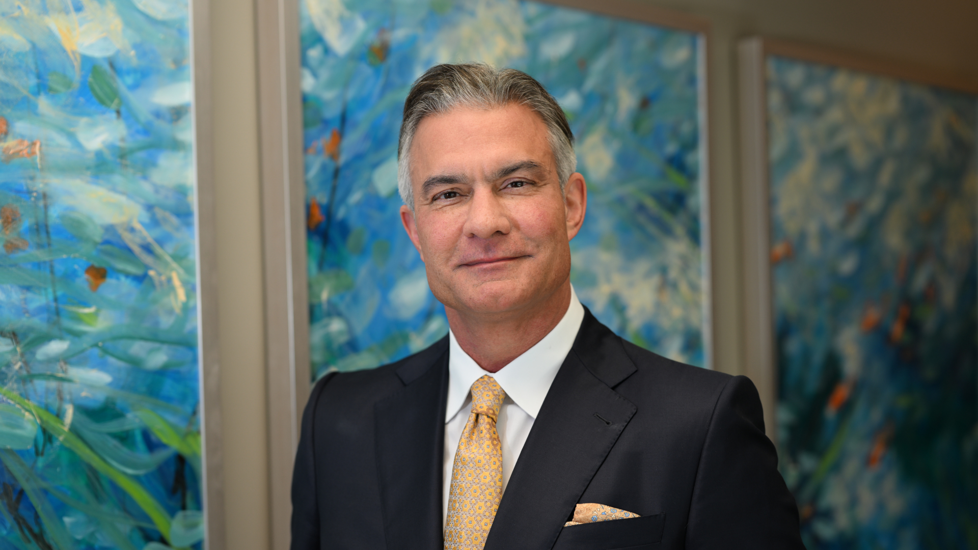 Man in a suit smiles, standing in front of abstract blue and white art.