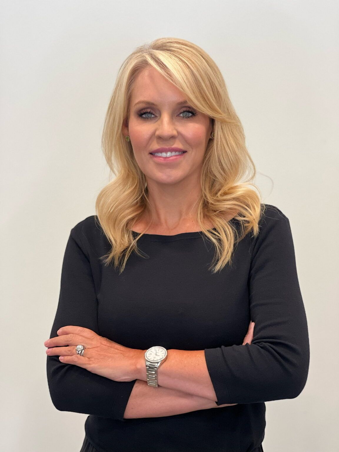Woman with blonde hair, arms crossed, wearing a black top and a watch, smiling, against a white backdrop.