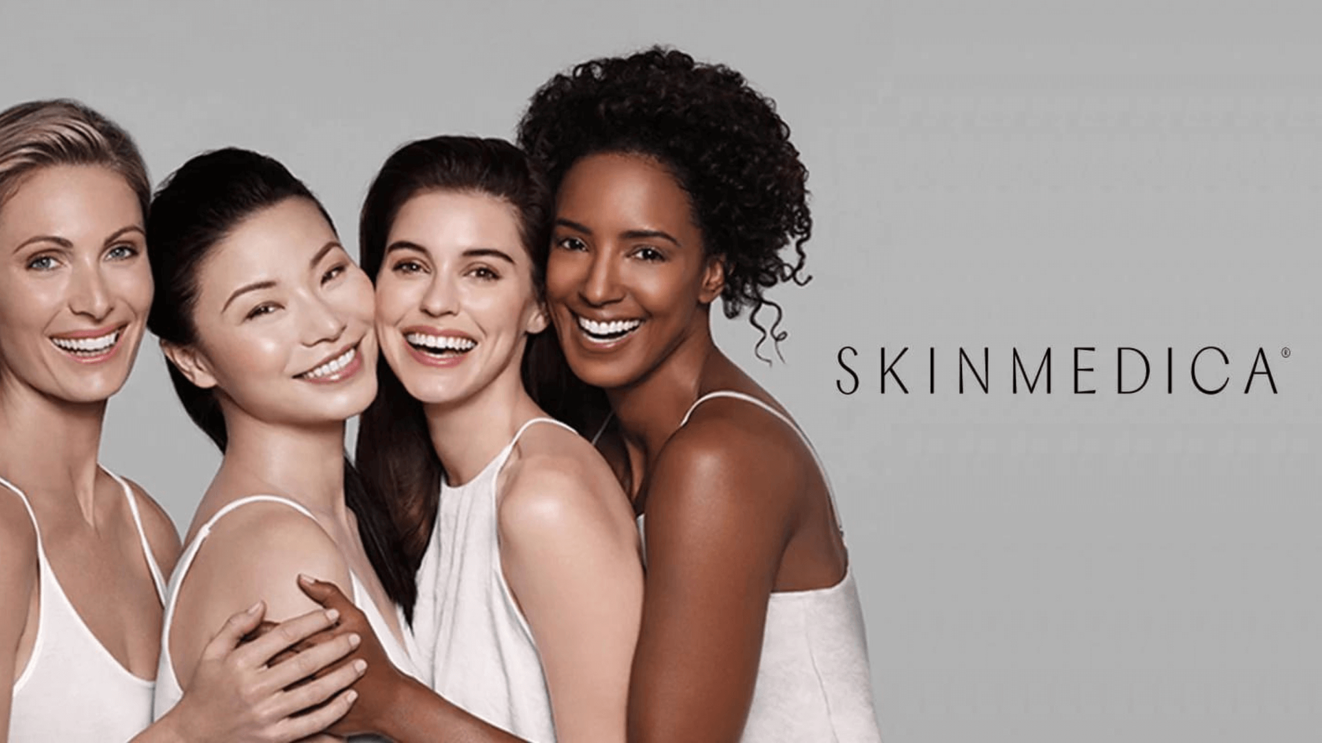 Four women of different skin tones smiling, embracing, against a gray background; 