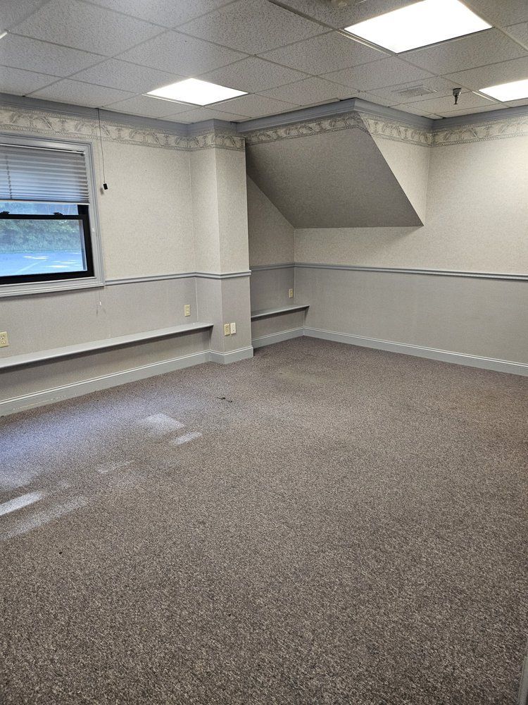 Empty carpeted office room with white walls, windows, fluorescent ceiling lights, and a built-in shelf niche.