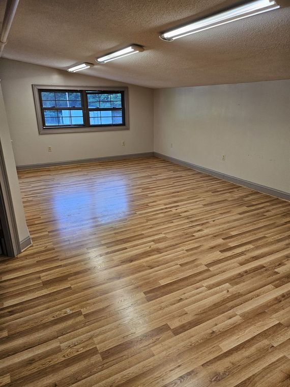 Empty room with wood floors, fluorescent lights, and a window along one wall.