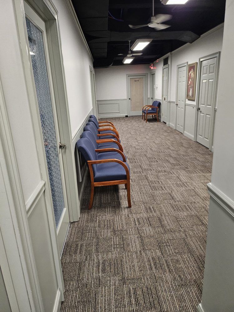 Quiet office hallway with blue chairs along the wall, patterned carpet, and closed doors.