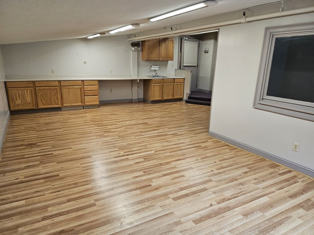 Empty basement room with wood-look flooring, cabinets along one wall, and a window on the right.