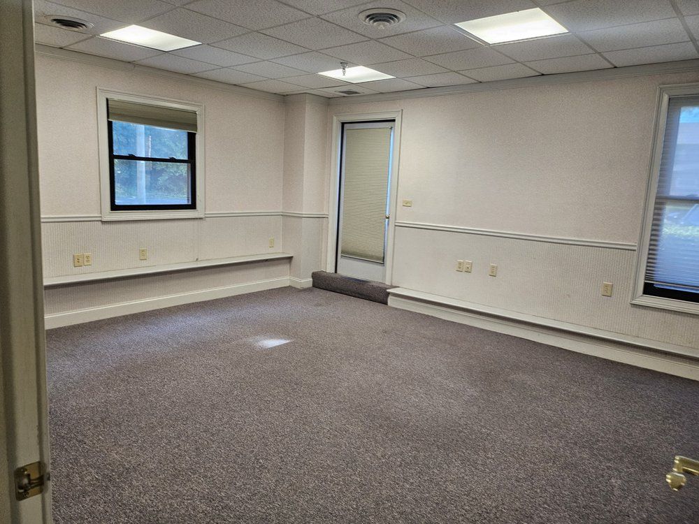 Empty office room with beige carpet, white walls, windows, and a central doorway.