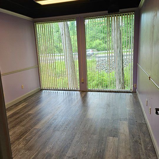 Empty room with gray wood floors, lavender walls, and sliding glass doors with vertical blinds.