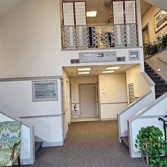 Bright lobby with staircase, balcony railing, carpeted floor, and chairs near the entrance