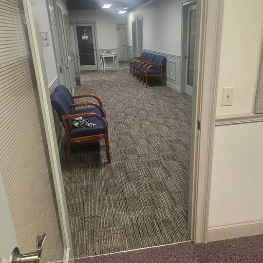 Empty office hallway with blue chairs, carpeted floor, and an open door in the foreground