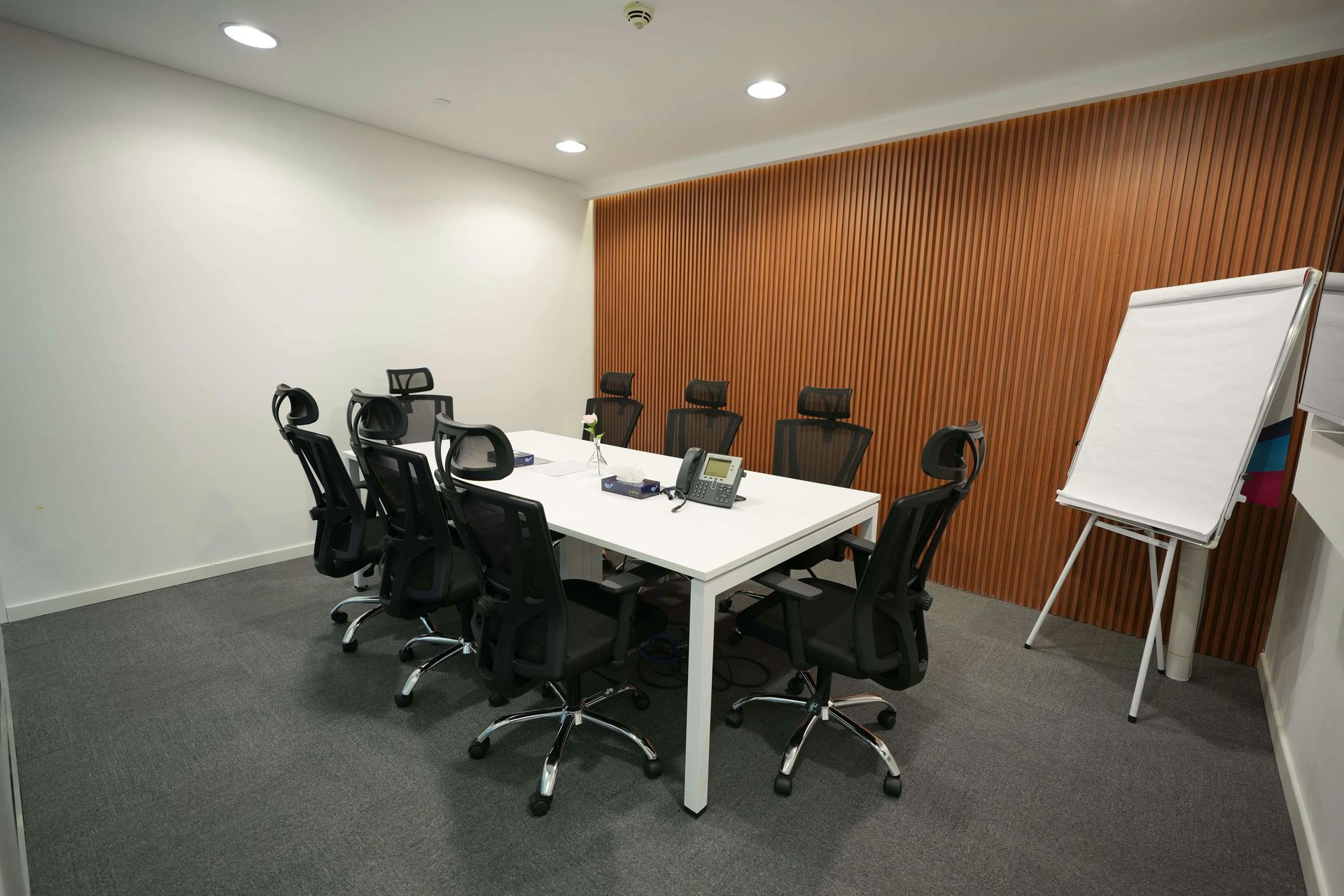 Conference room with white table, black chairs, TV, and flip chart beside wooden accent wall.