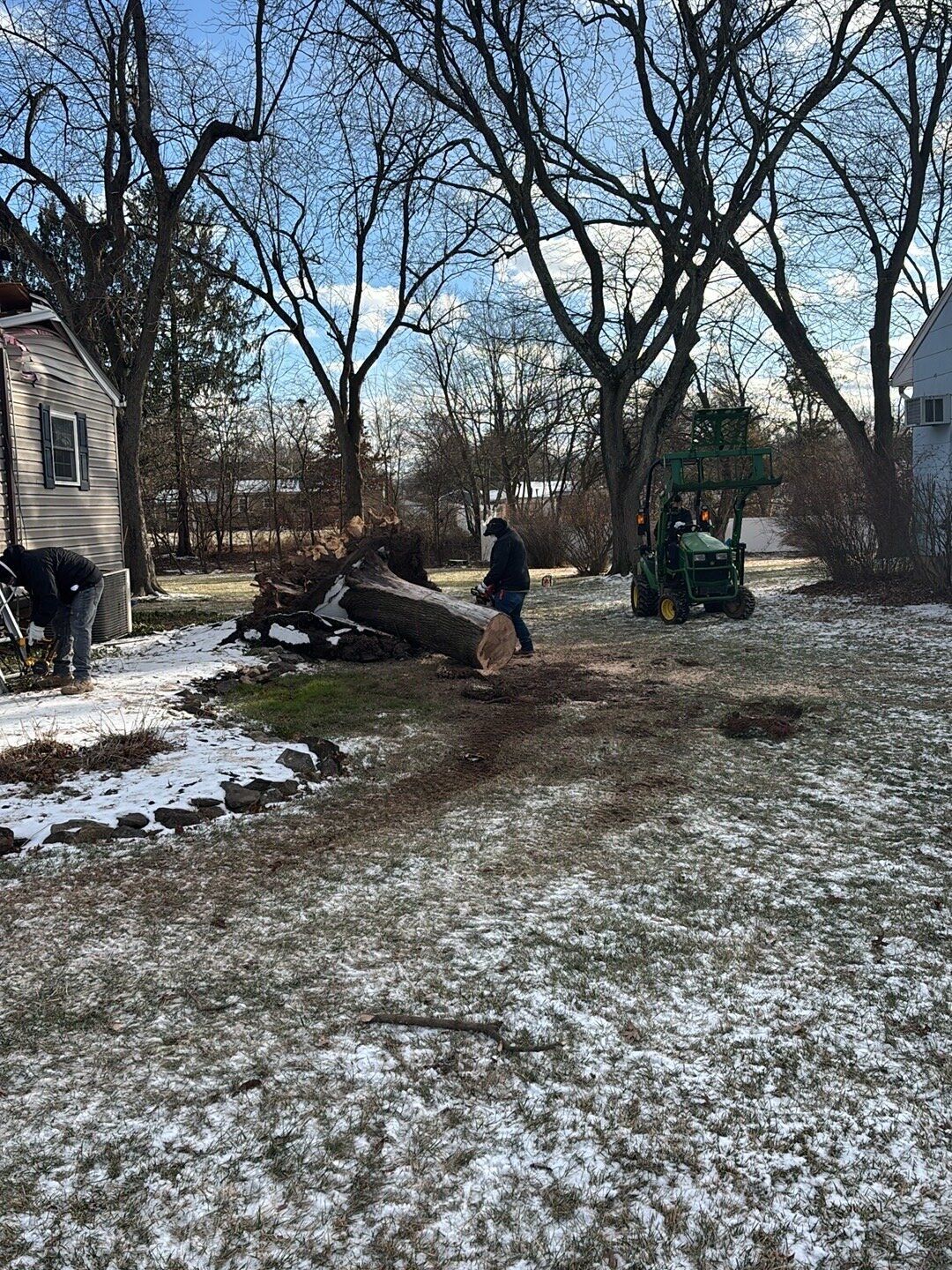Crew member using a chainsaw on a fallen tree trunk in a snowy backyard while a tractor stands ready