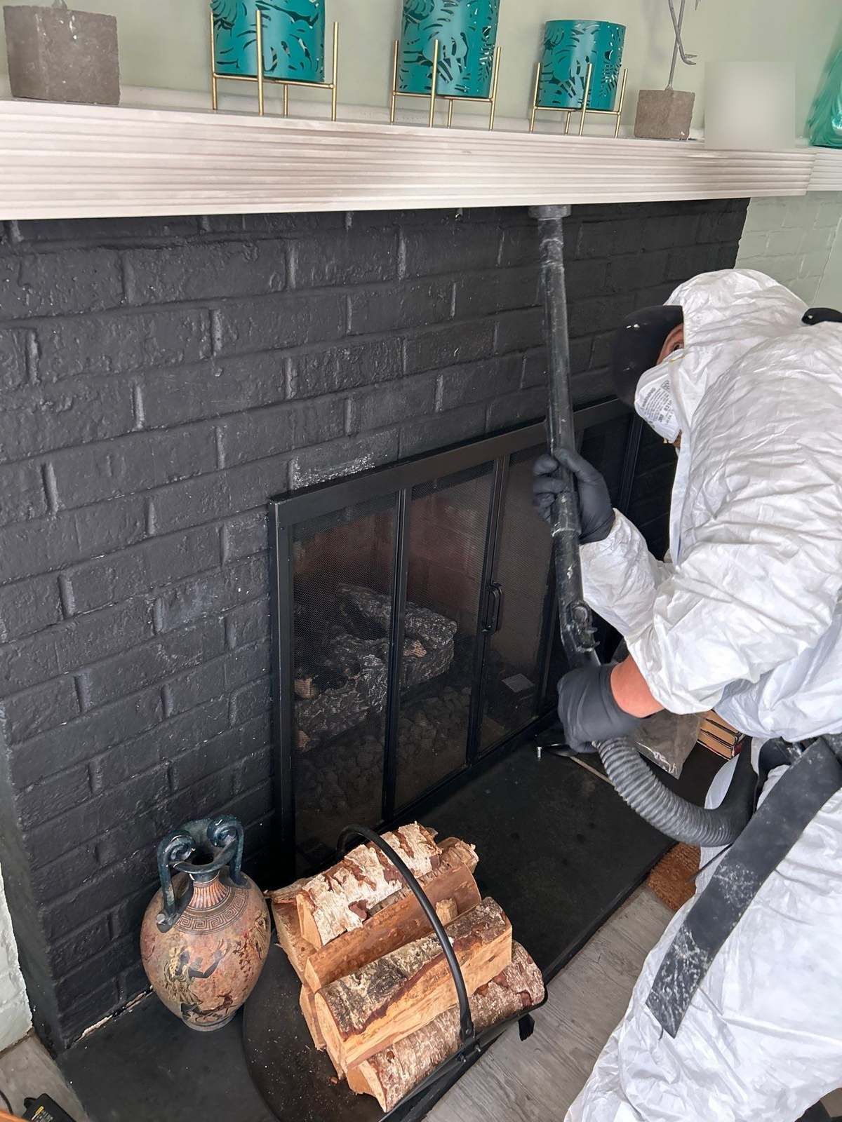 Technician in a protective suit and respirator vacuuming soot from a painted brick fireplace and its doors