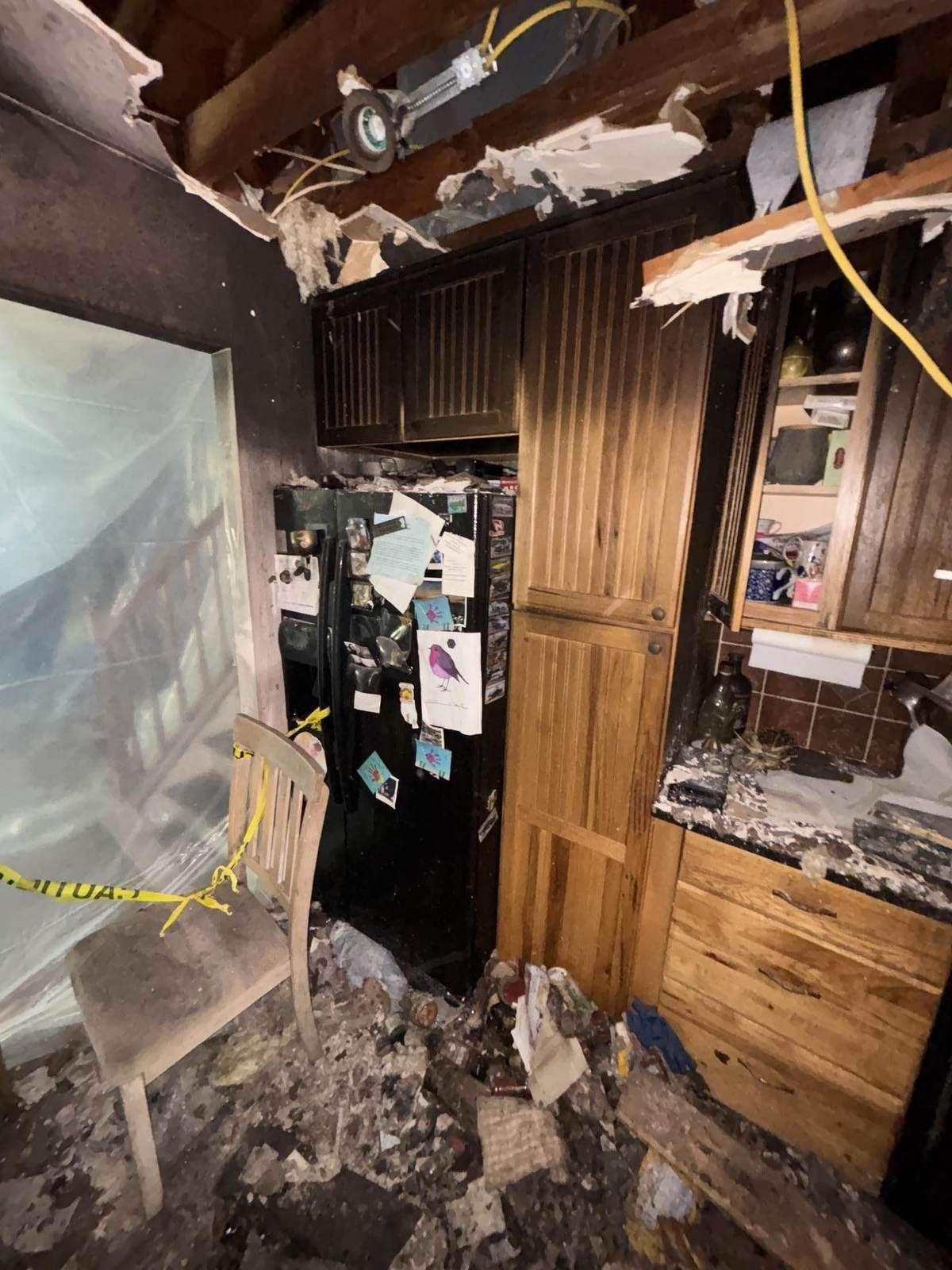 Soot-stained kitchen pantry cabinets and refrigerator under an opened fire-damaged ceiling