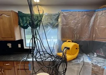 Kitchen with cabinets and window, draped in plastic. A yellow air mover and tangled cords are present.