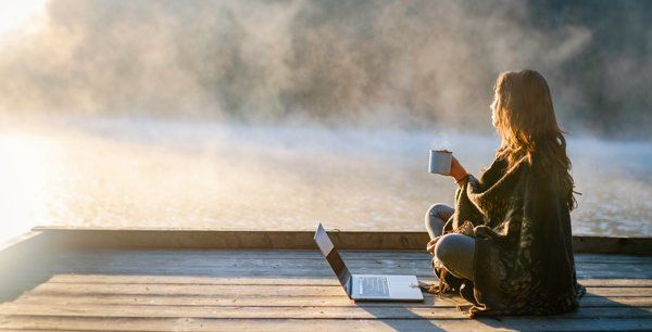 A woman is sitting on a dock with a laptop and a cup of coffee.