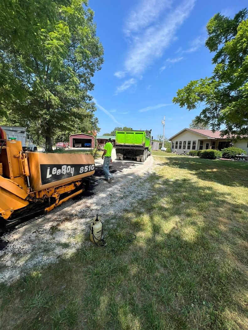 Construction equipment and dump truck on a gravel driveway beside houses under a blue sky