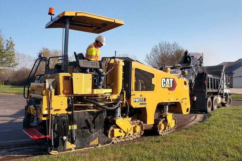 Yellow road paver on roadside with worker in safety gear, laying asphalt near parked truck