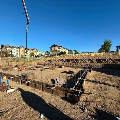 Residential construction site with concrete foundations, rebar, and houses in the background under a clear blue sky