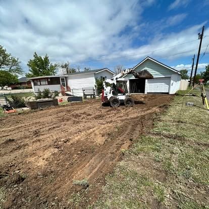 Worker on a skid steer grading a dirt yard in front of a house and garage under a cloudy sky