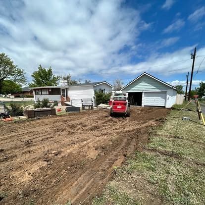 Red skid steer on a dirt driveway beside a light blue garage and a mobile home under a partly cloudy sky