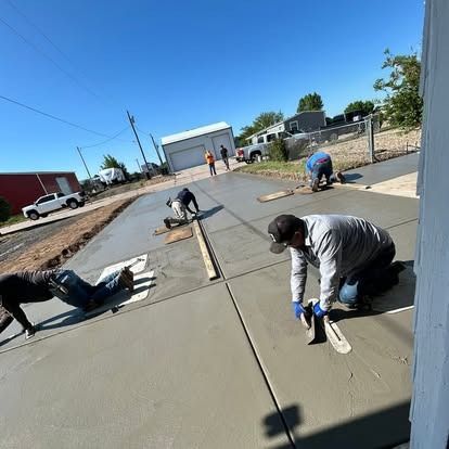 Workers smoothing fresh concrete on a road under bright blue sky, with houses and vehicles nearby